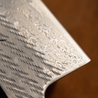 Close-up of a silver patterned steel knife blade with a hammered texture and a black signature, set against a dark brown background.