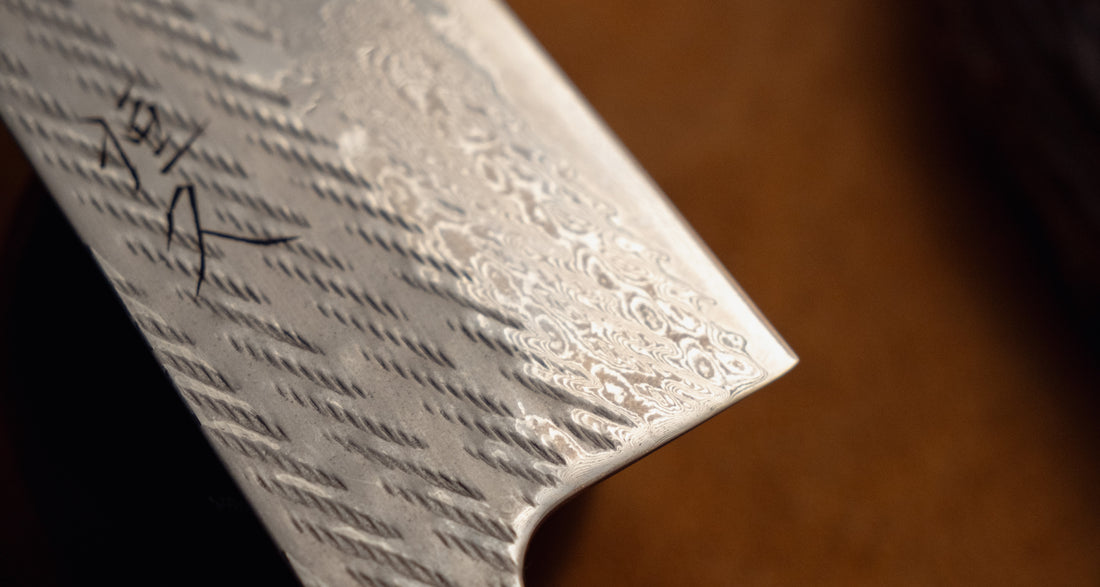 Close-up of a silver patterned steel knife blade with a hammered texture and a black signature, set against a dark brown background.