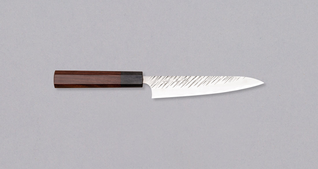 Kurosaki Petty Fujin is a uniquely looking blade from the talented Yu Kurosaki. Petty is a small kitchen knife perfect for all those delicate tasks where a larger knife is deemed unwieldy. Core from SG2 powder steel is enveloped in softer steel with a striking tsuchime pattern. Fitted with an octagonal rosewood handle.