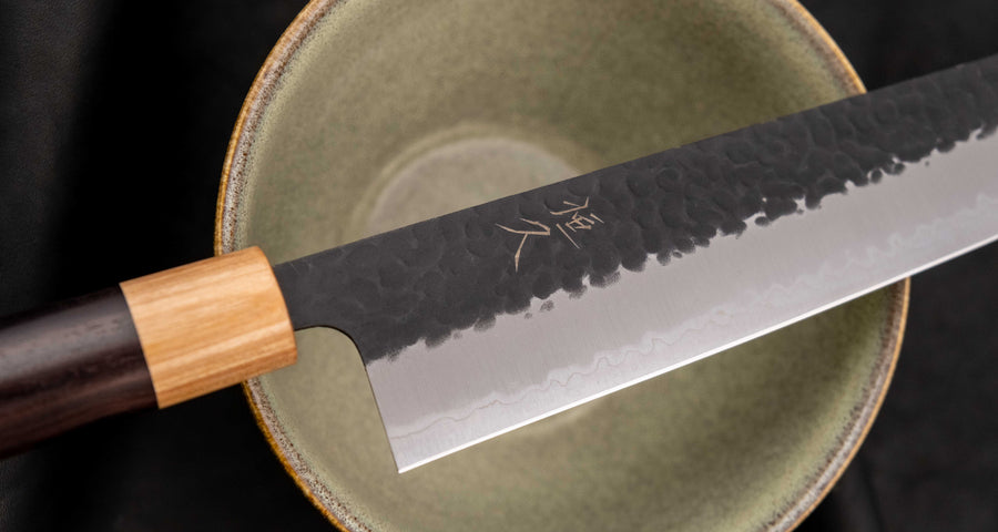 Tsunehisa Kiritsuke AS Kuro-uchi 240mm (9.4") is a multi-purpose knife with a black Kuro-uchi protective patina and tsuchime hammer indentations. Its length makes it great for cutting larger pieces of meat and fish. The Kuro-uchi (black) patina on the upper part of the knife gives it a unique and rustic appearance.