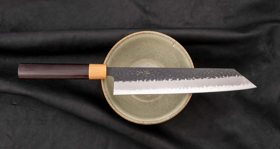 Tsunehisa Kiritsuke AS Kuro-uchi 240mm (9.4") is a multi-purpose knife with a black Kuro-uchi protective patina and tsuchime hammer indentations. Its length makes it great for cutting larger pieces of meat and fish. The Kuro-uchi (black) patina on the upper part of the knife gives it a unique and rustic appearance.